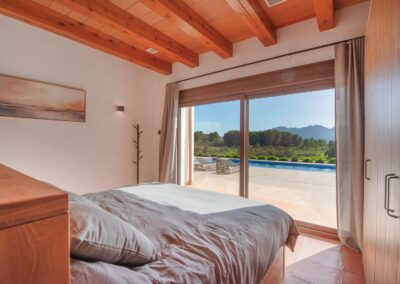 Habitación con suelo de terracota y techo de vigas de madera, con una cama orientada hacia unas puertas acristaladas que se abren a un jardín y un paisaje montañoso