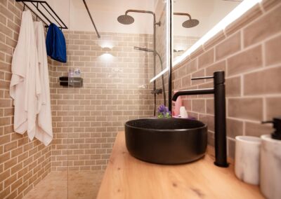 Cuarto de baño con un lavabo negro, inodoro y paredes de azulejos. Al fondo el espacio de la regadera tipo lluvia