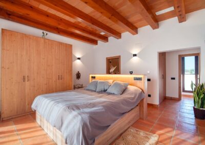 Habitación de estilo rústico-moderno inundada de luz solar, con suelo de terracota, techo de vigas y una cama con cabecero iluminado. Un amplio armario de madera está a su lado