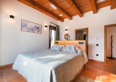 Habitación de estilo rústico-moderno inundada de luz solar, con suelo de terracota, techo de vigas y una cama con cabecero iluminado. Un cuadro de colores cuelga en la pared.