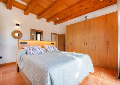 Habitación de estilo rústico con un gran armario de pino a la derecha de la cama. El cabecero de la cama tiene una luz integrada y el techo es de vigas de madera.