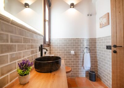 Aseo con azulejos, un lavabo negro sobre un mueble de madera y suelo de terracota.