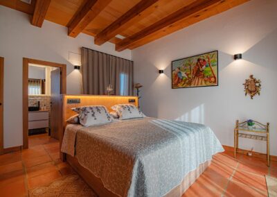 Panorámica de un dormitorio que combina suelo de terracota y techo de vigas de madera con mobiliario moderno. A la derecha hay un cuadro, y en el centro, una cama con cabecero de luz integrada. Al fondo se ve la entrada a un baño en suite