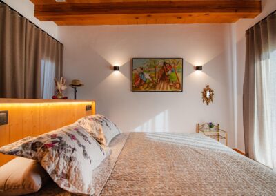 Amplia habitación de estilo rústico con un cuadro decorativo, una cama con cabecero iluminado y un techo de vigas de madera a juego."