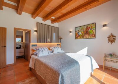 Dormitorio soleado con cama de matrimonio, techo con vigas de madera y acceso a un baño.