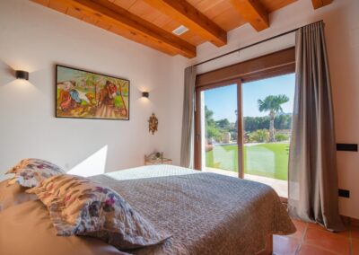 Habitación con suelo de terracota y techo de vigas de madera, con una cama orientada hacia unas puertas acristaladas que se abren a un jardín y un paisaje montañoso