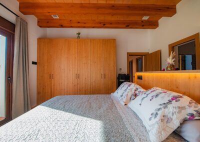 Vista desde la cama de un dormitorio, enfocando el cabecero de madera con luz integrada, el closet moderno y las vigas de madera del techo.