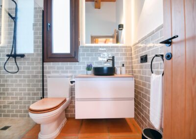 Baño con paredes de azulejo gris, un lavabo redondo y negro sobre un mueble de madera, y suelo de baldosas de terracota.