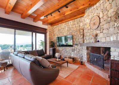 Amplia sala de estar con un sofá en forma de L frente a una pared de piedra que integra una chimenea y un televisor. El techo tiene vigas de madera y grandes ventanales dan al exterior
