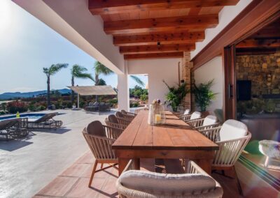 Un amplio porche con techo de vigas de madera y un comedor al aire libre y la piscina a una lado junto a las tumbonas
