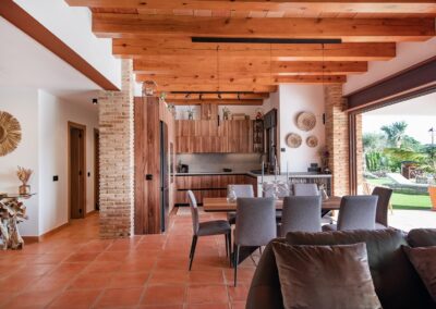 Interior de un salón de planta abierta con vigas de madera, suelo de terracota y una cocina moderna al fondo