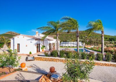 Vista exterior de una finca con una casa blanca y tejado de tejas, rodeada de un jardín joven con palmeras y con montañas al fondo bajo un cielo despejado.